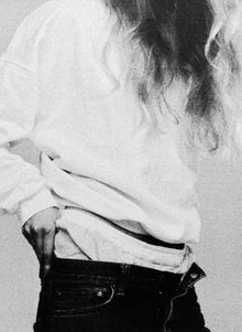 A white sweatshirt lifted at the waist over unbuttoned dark jeans, close on the hip.